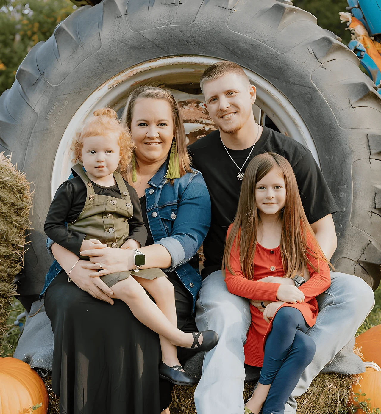 Cassie Large, owner of Gracie Jane on Main, smiling with her husband and two young daughters while sitting on hay bales in front of a tractor wheel during a fall family photo shoot in Michigan