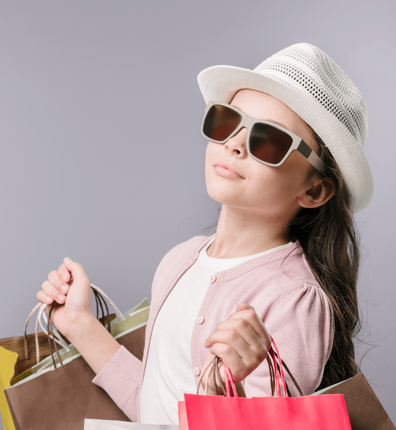 Stylish young girl wearing sunglasses, a white hat, and pink cardigan while holding shopping bags