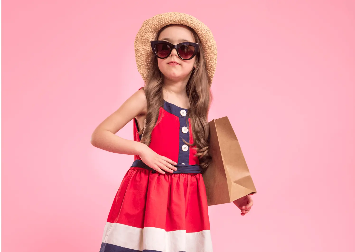 Stylish young girl in a red, white, and navy summer dress wearing sunglasses and a straw hat while holding a shopping bag against a pink background.