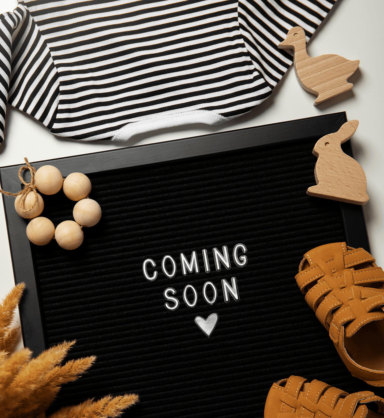 Flat lay of baby outfit, wooden toys, and sandals surrounding a black letter board with 'Coming Soon' text